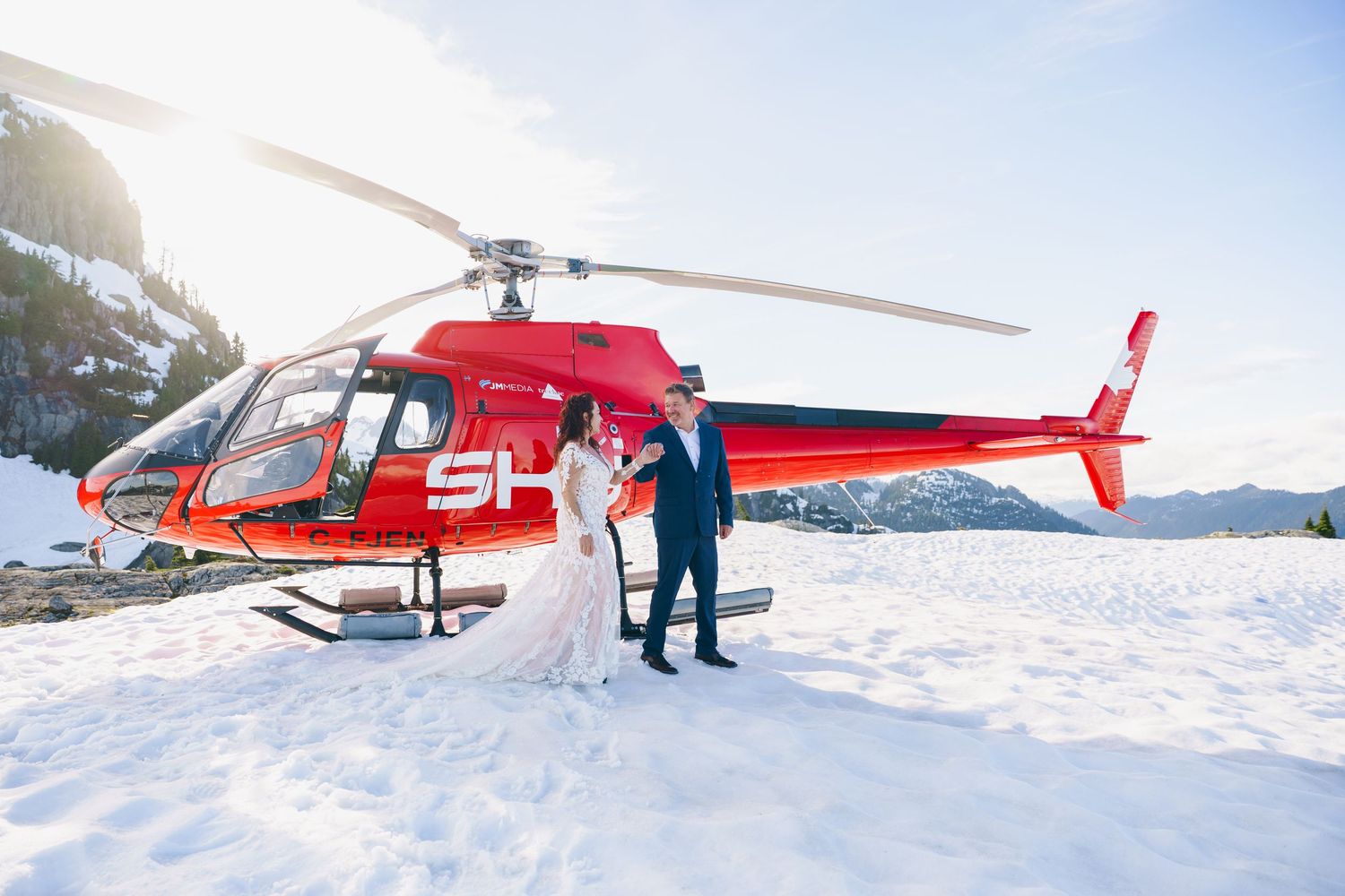 Red helicopter parked on snowy mountain terrain with passengers and crew preparing for takeoff.
