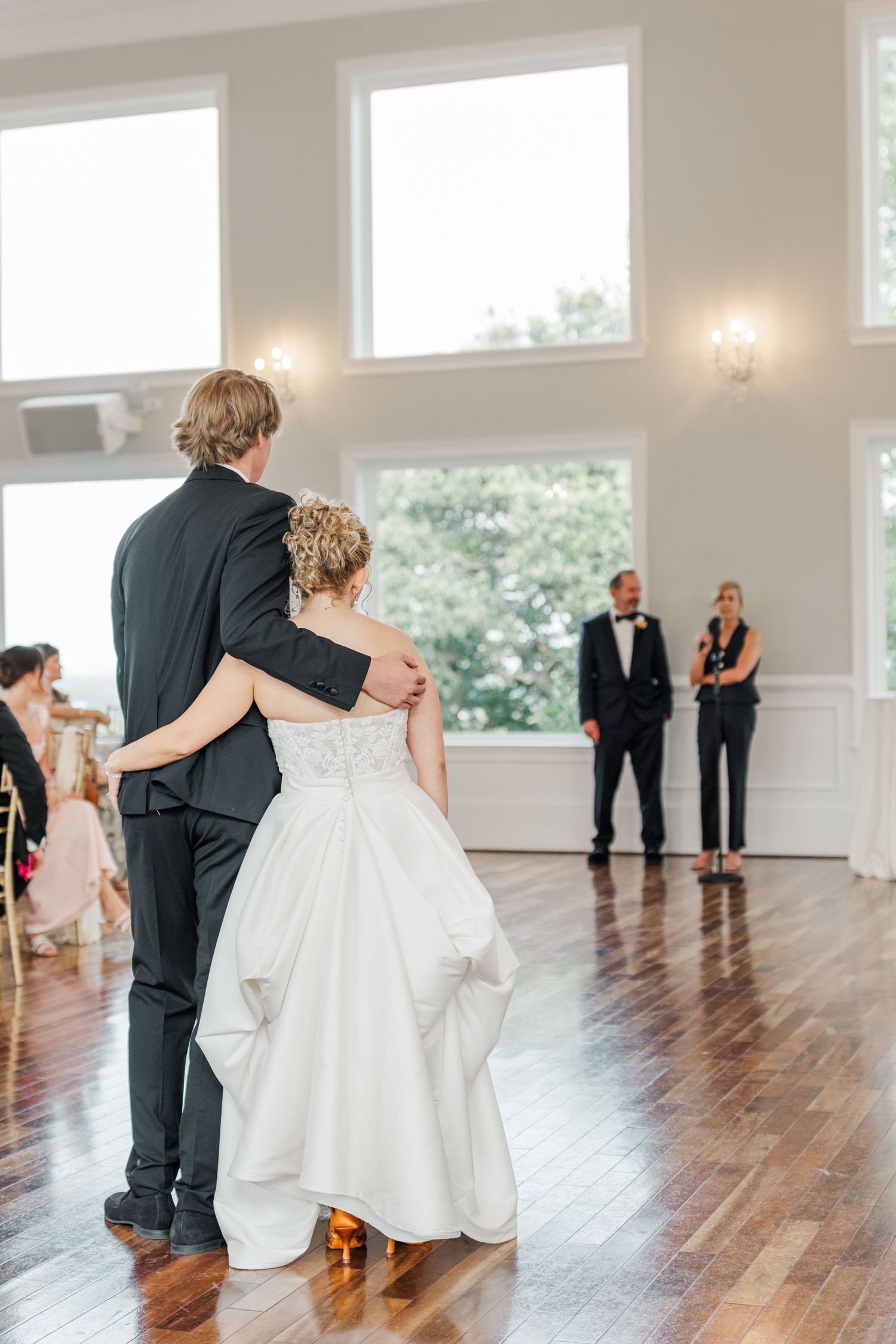 Couple embraces while dancing in an elegant white ballroom with large windows.