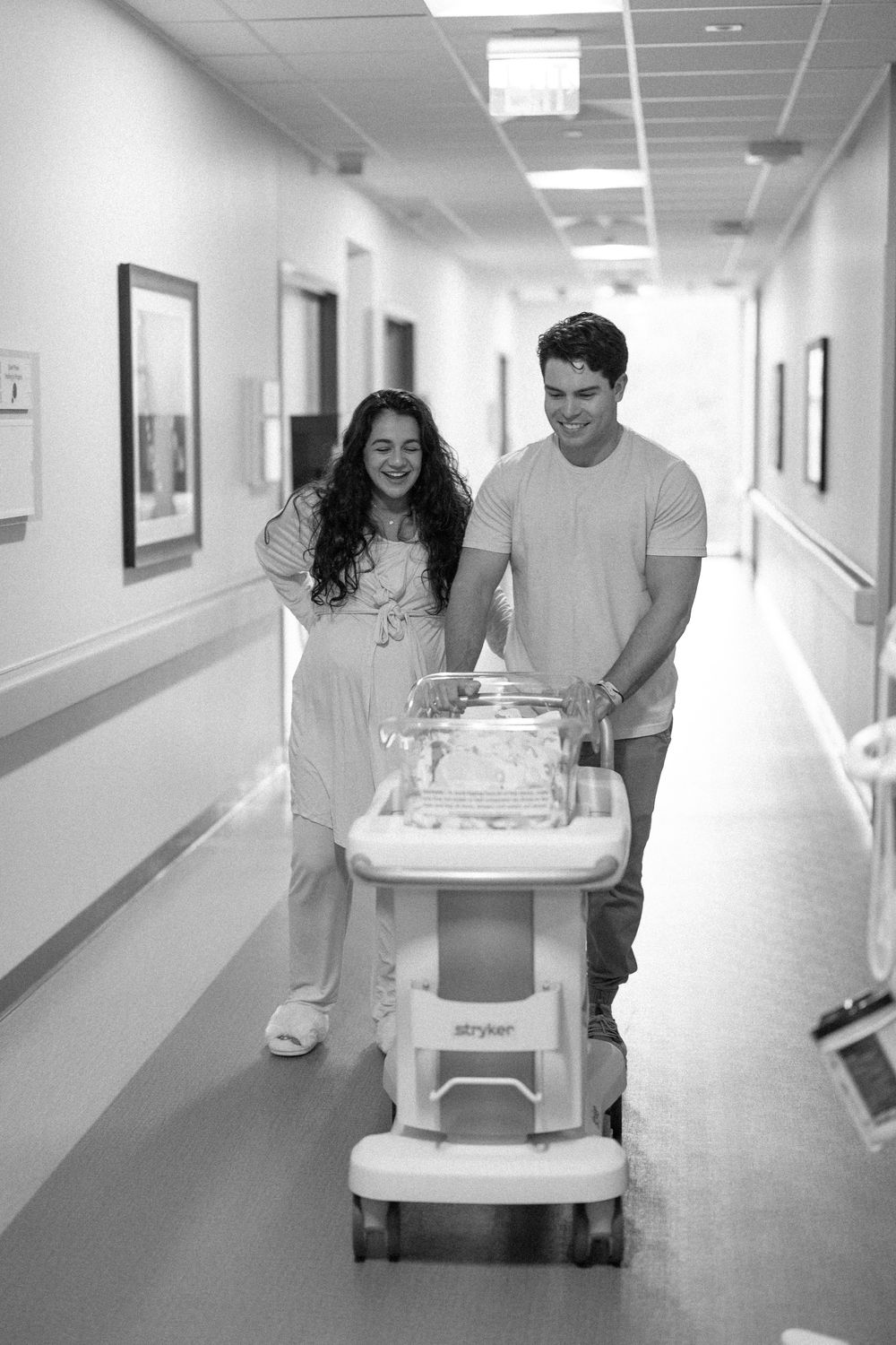 A couple walks down a hospital hallway pushing a bassinet cart, captured in black and white photography.