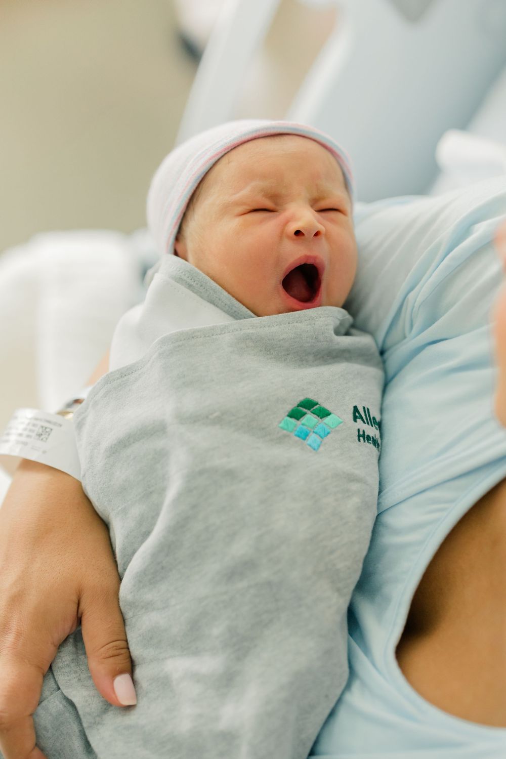 A newborn baby wrapped in a light gray swaddle smiles and yawns while lying in a hospital bassinet.
