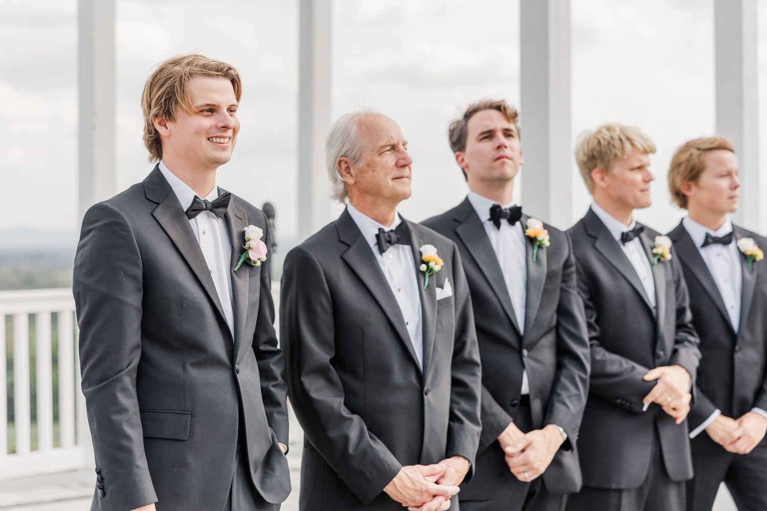 Groomsmen in black tuxedos lined up at outdoor wedding ceremony.