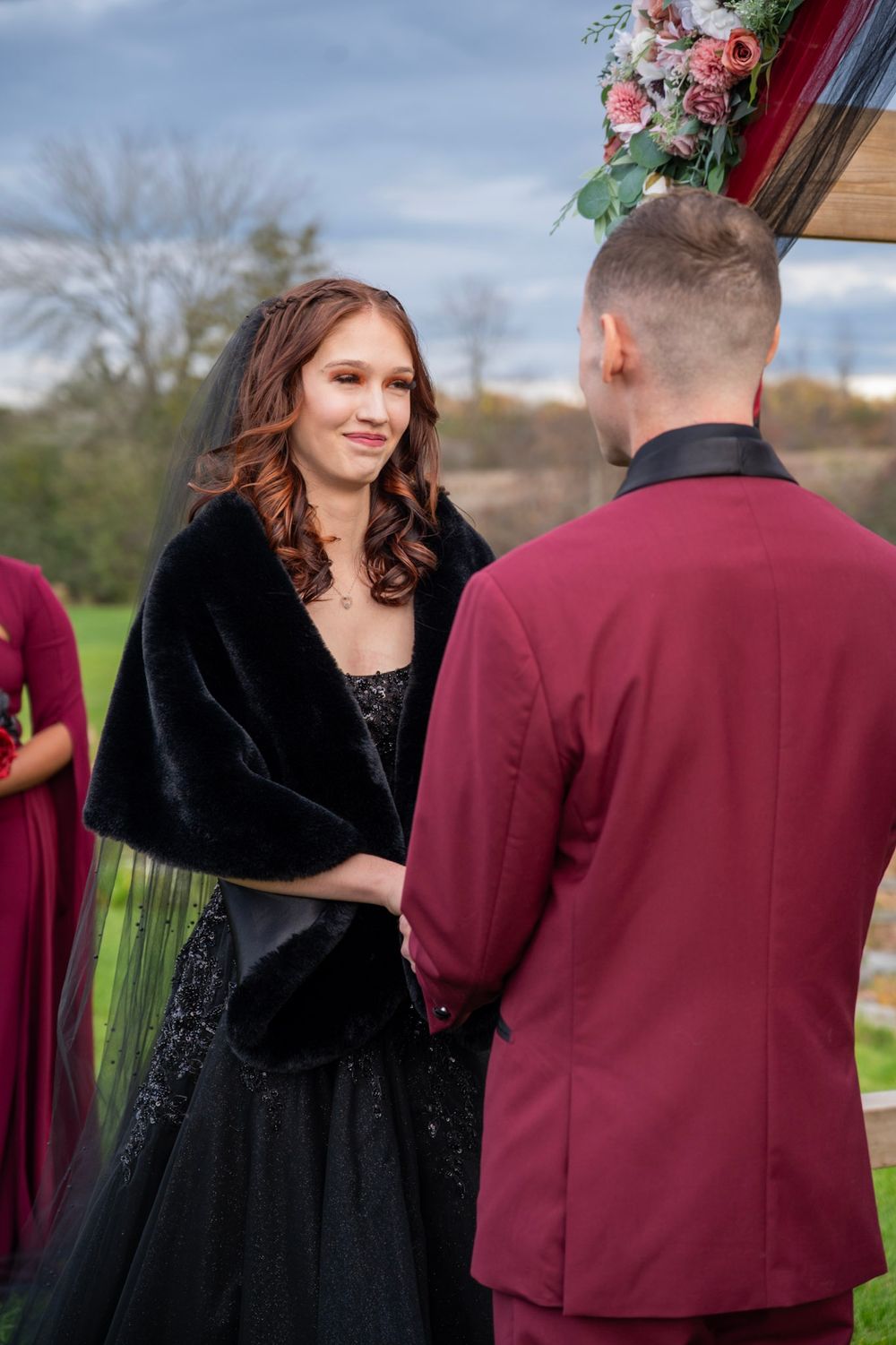 Bride in black wedding dress and fur shawl smiles at groom during outdoor wedding ceremony.