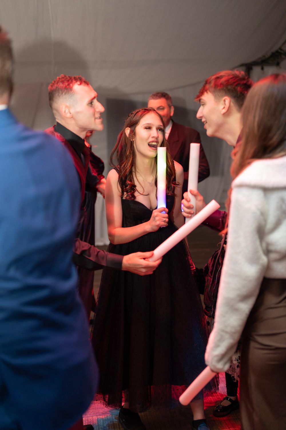 Bride in black dress laughing with guests holding glow sticks on wedding reception dance floor.