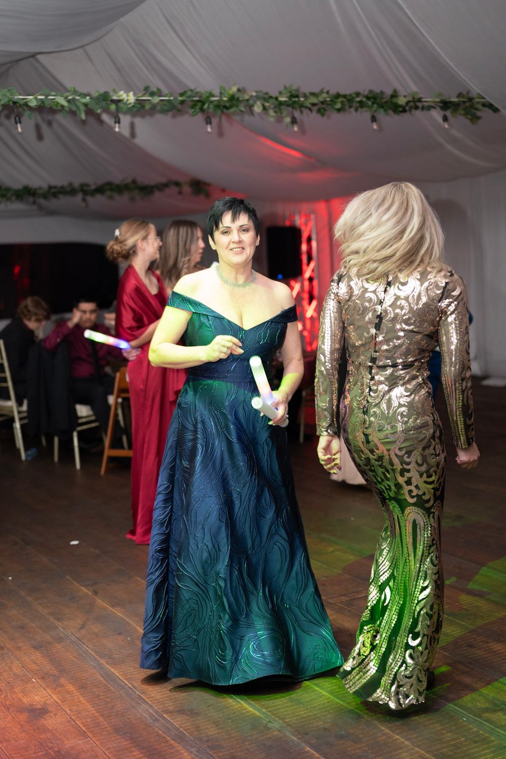 Two elegantly dressed women in green peacock gown and gold sequin dress dancing at wedding reception.