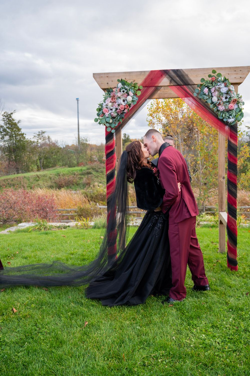 Bride in black gown with veil and groom in burgundy suit share first kiss under decorated wooden arch outdoors.