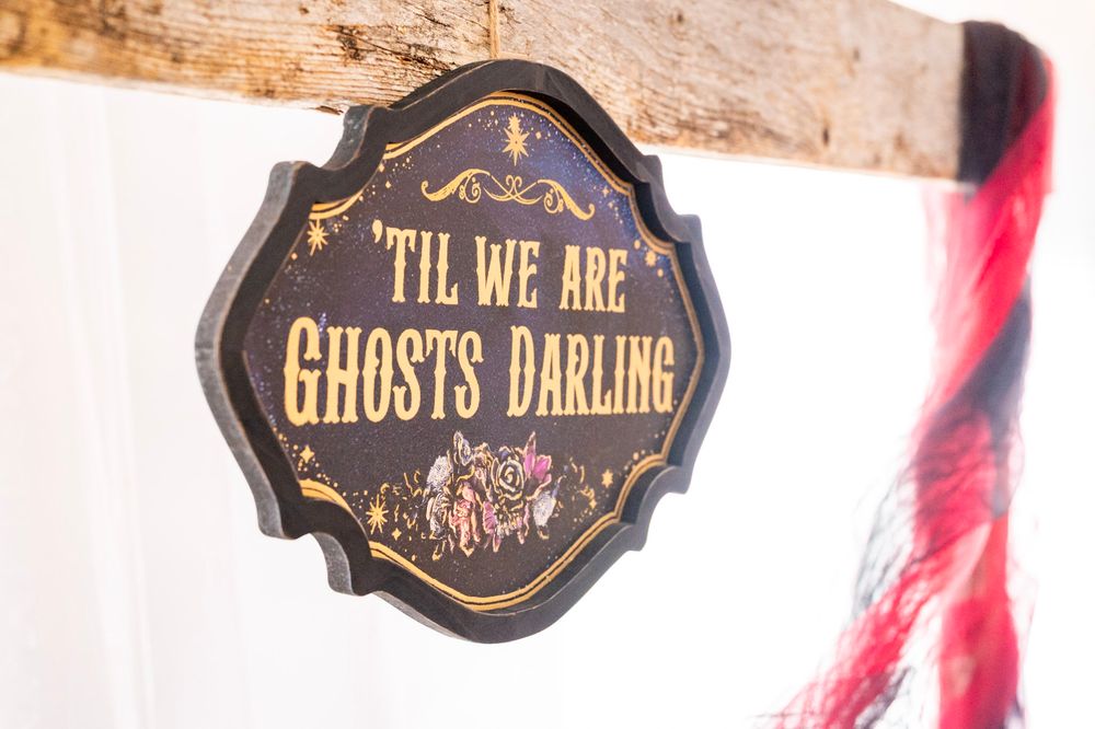 Black decorative sign reading 'Til We Are Ghosts Darling' with floral accents hanging from a wooden beam.