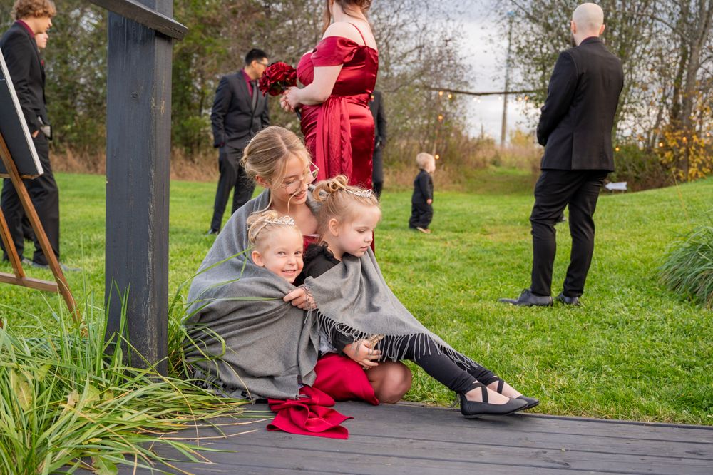 Woman sitting on deck cuddles two young children in gray outfits during outdoor wedding celebration.