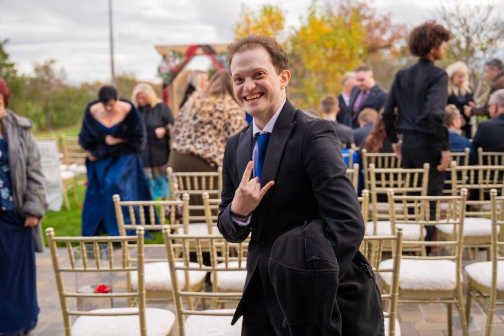 Smiling wedding guest in black suit making a rock hand gesture at an outdoor ceremony.