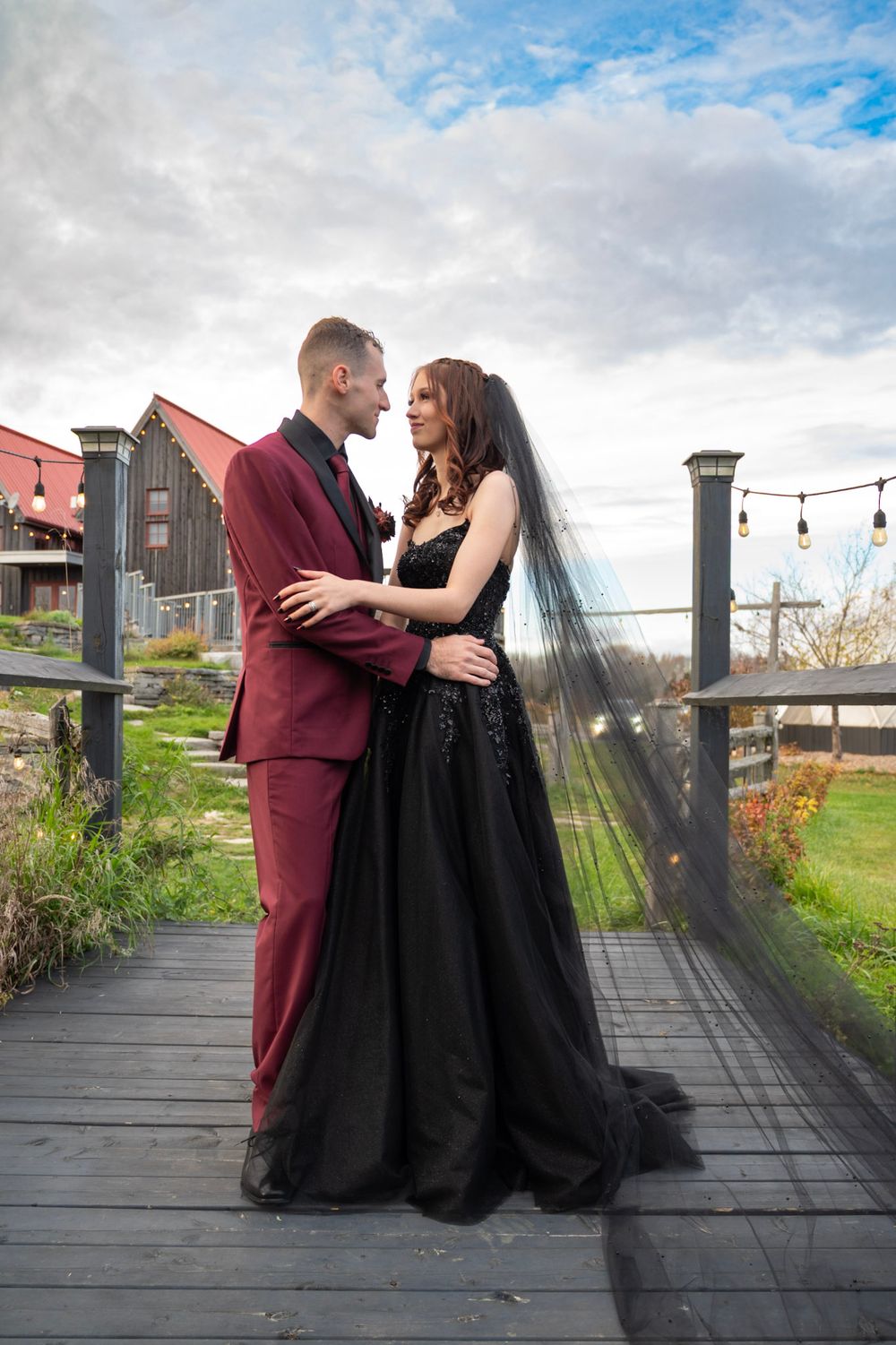 Bride in black gown with veil and groom in burgundy suit embrace on a wooden bridge.