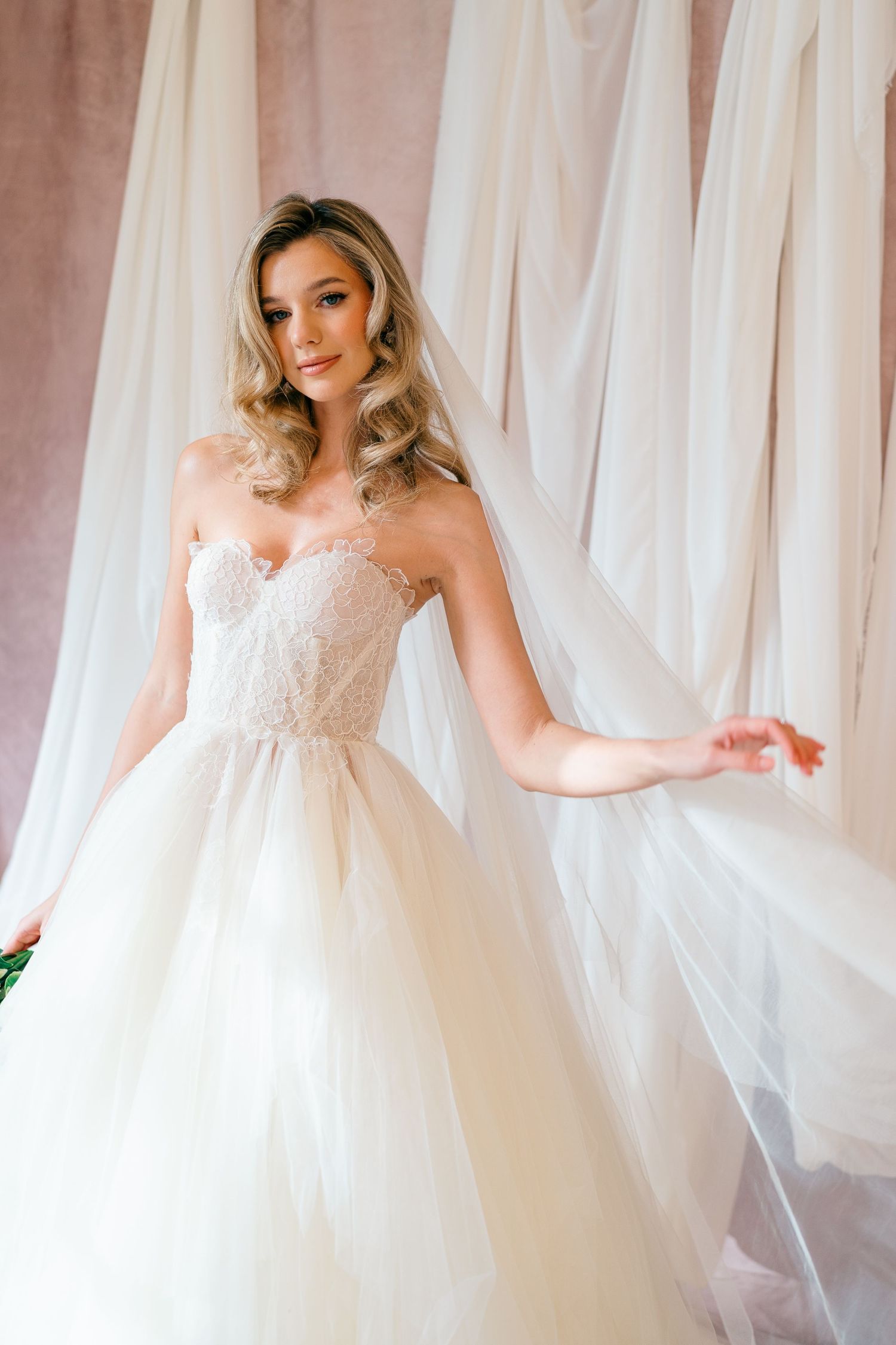 A bride in a strapless lace ballgown poses elegantly against flowing white curtains in a romantic bridal portrait.