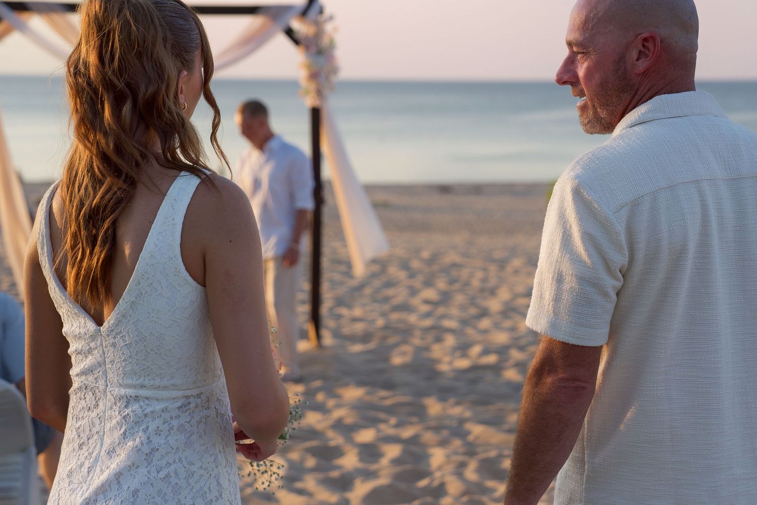 A couple walks hand in hand on a sandy beach at sunset wearing white outfits.