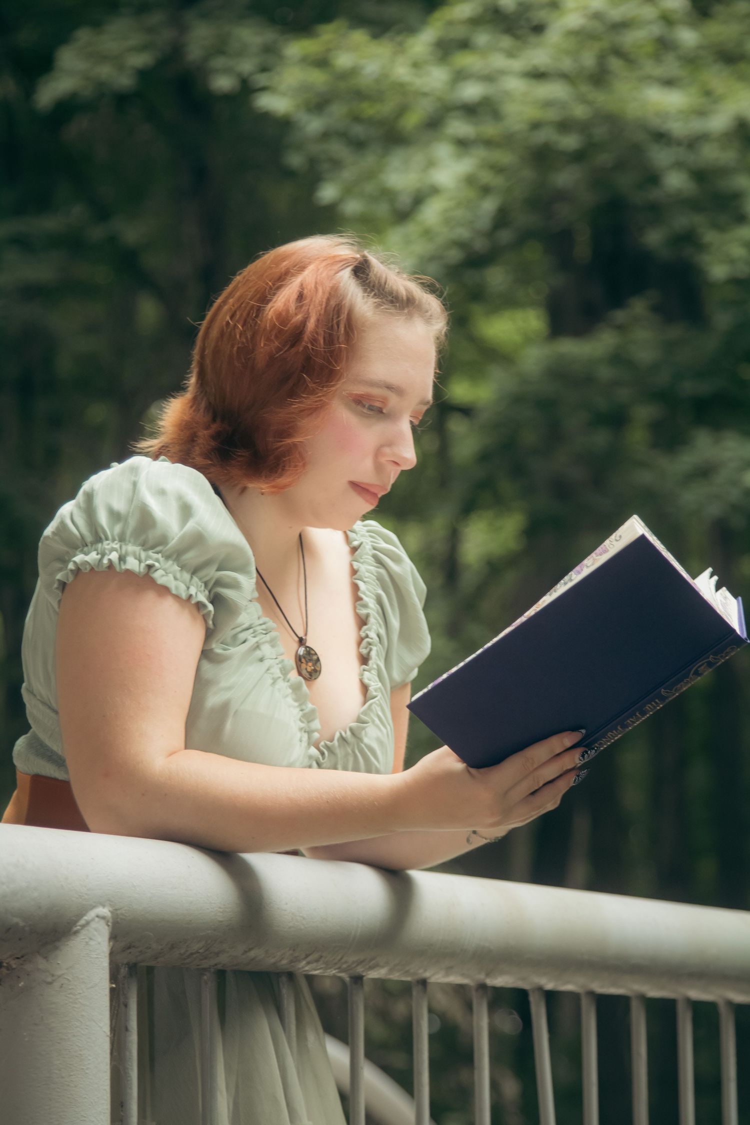 A person in a vintage dress reads a book while leaning on a white railing outdoors with trees in the background.