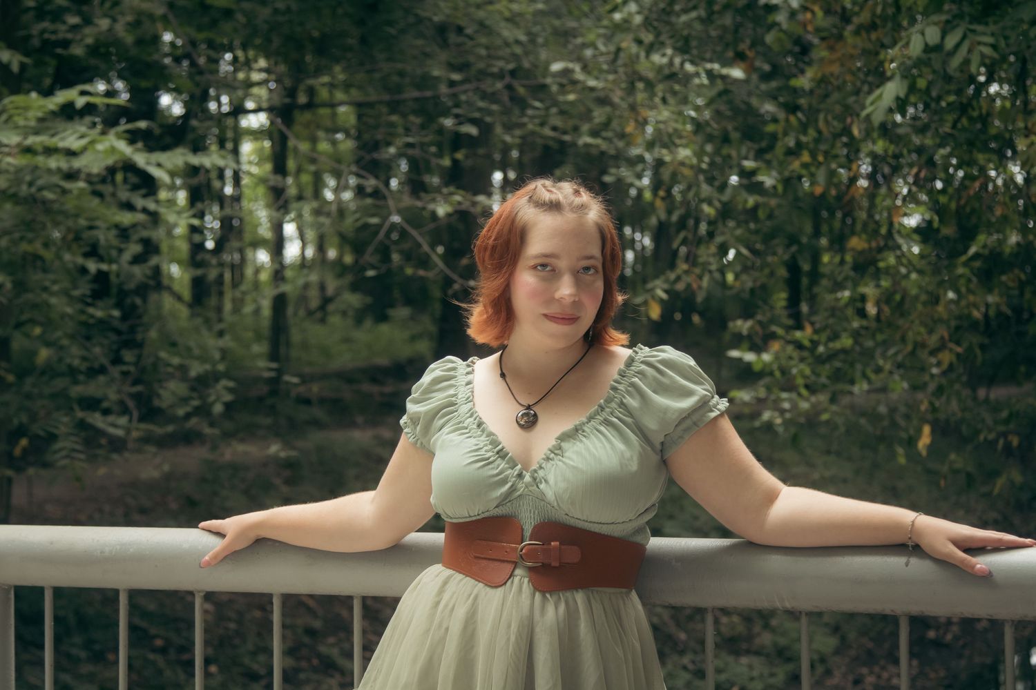 A person in a vintage-style dress poses outdoors in a natural setting with trees and foliage in the background.