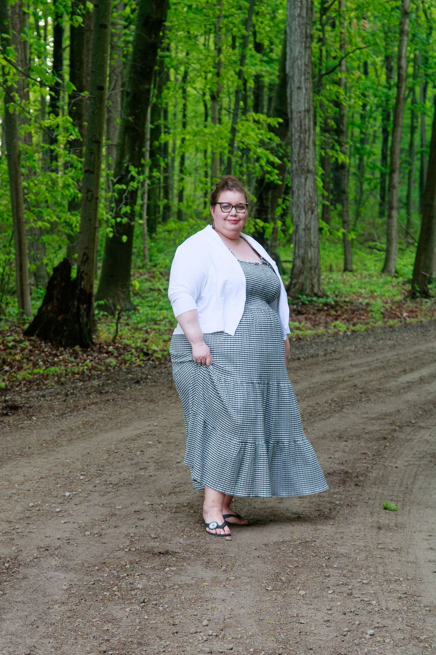 A plus-size woman in a gray maxi dress and white cardigan stands on a dirt path surrounded by lush green forest trees.
