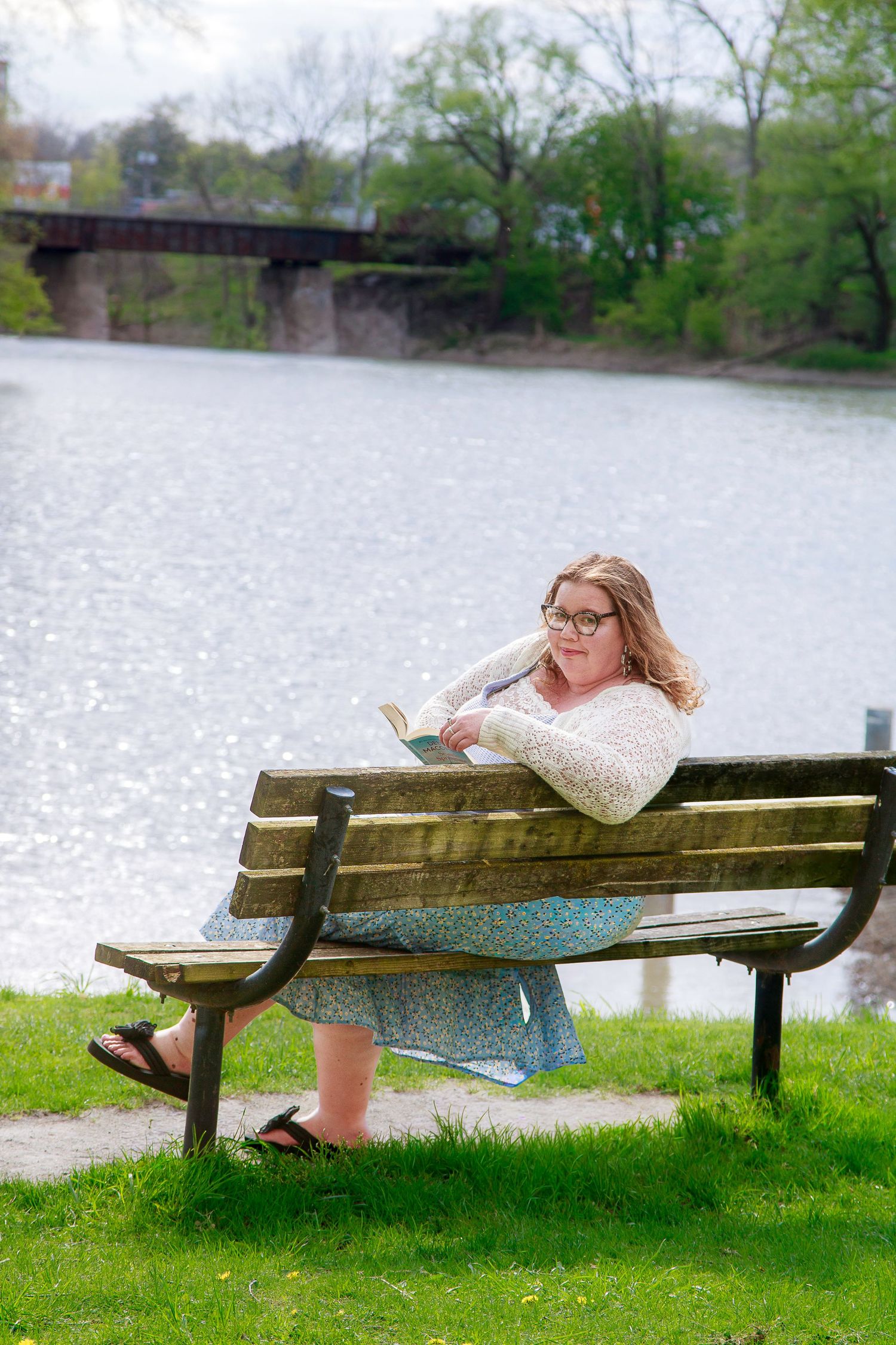 Woman in glasses reads book on wooden park bench beside a serene river, wearing a floral skirt and white cardigan.