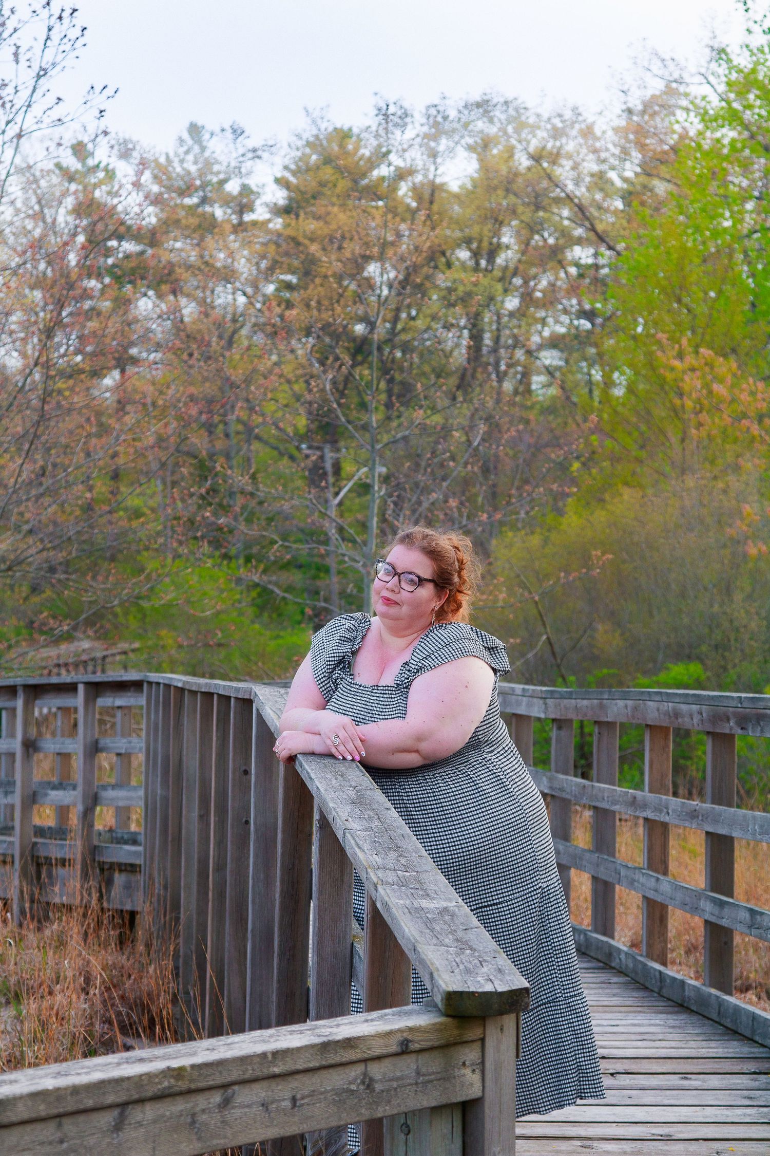 A plus-size woman in a gray dress leans on a wooden bridge railing, smiling outdoors amid autumn trees.