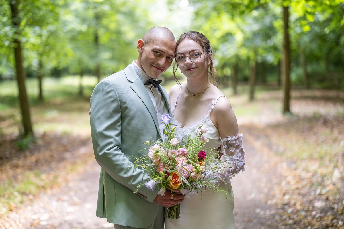 A couple in wedding attire pose together on a wooded path, the bride holding a vibrant wildflower bouquet.