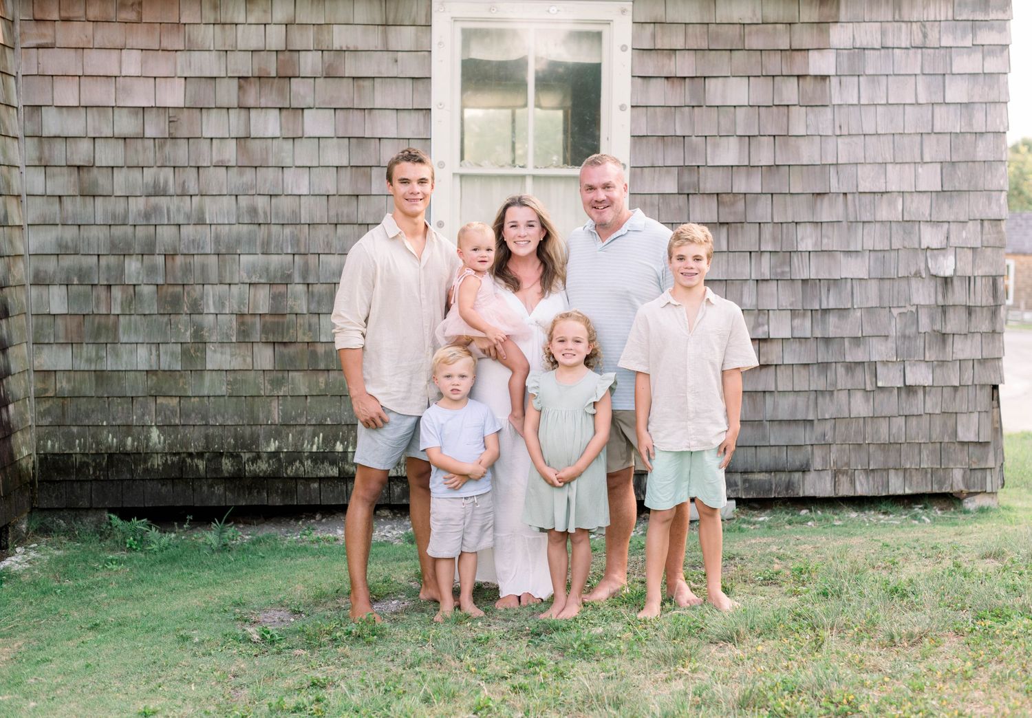A family of five stands together against a rustic shingled house exterior during a casual summer photoshoot.