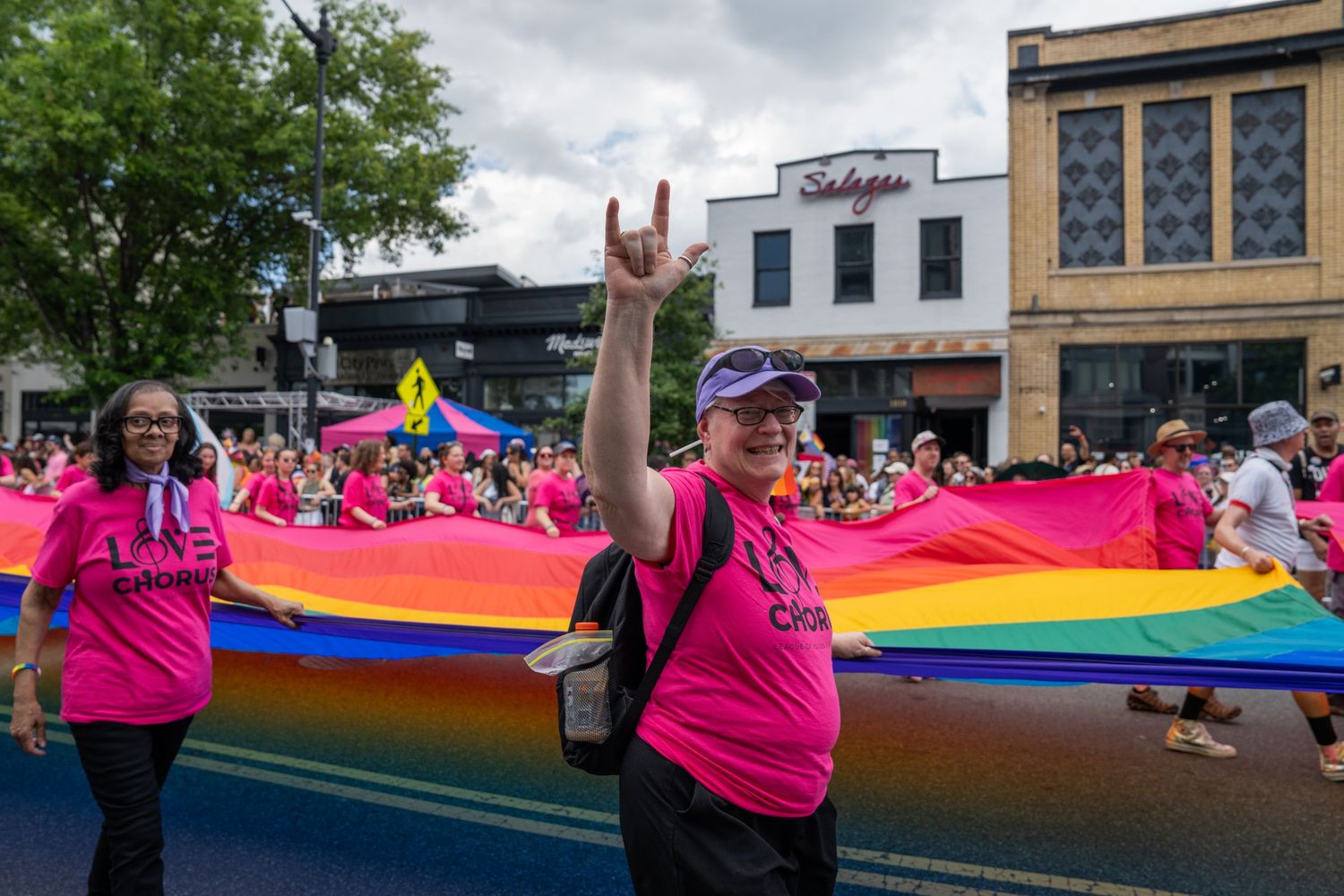 World Pride Parade: person walks by in a pink choir shirt carrying a part of a massive rainbow flag and doing the ASL sign for "I love you"