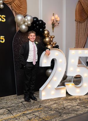 Young boy in black suit and pink tie leaning on illuminated number 25 at graduation event.