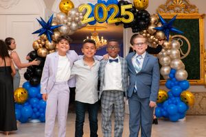 Four young boys in formal suits posing together under a 2025 graduation balloon arch.