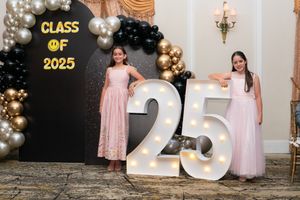 Two girls in pink dresses posing beside illuminated Class of 2025 marquee number sign.