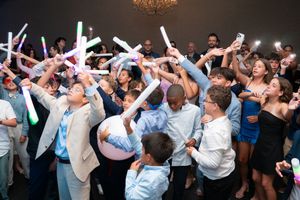 Excited crowd of children and adults raising colorful glow sticks at graduation party.