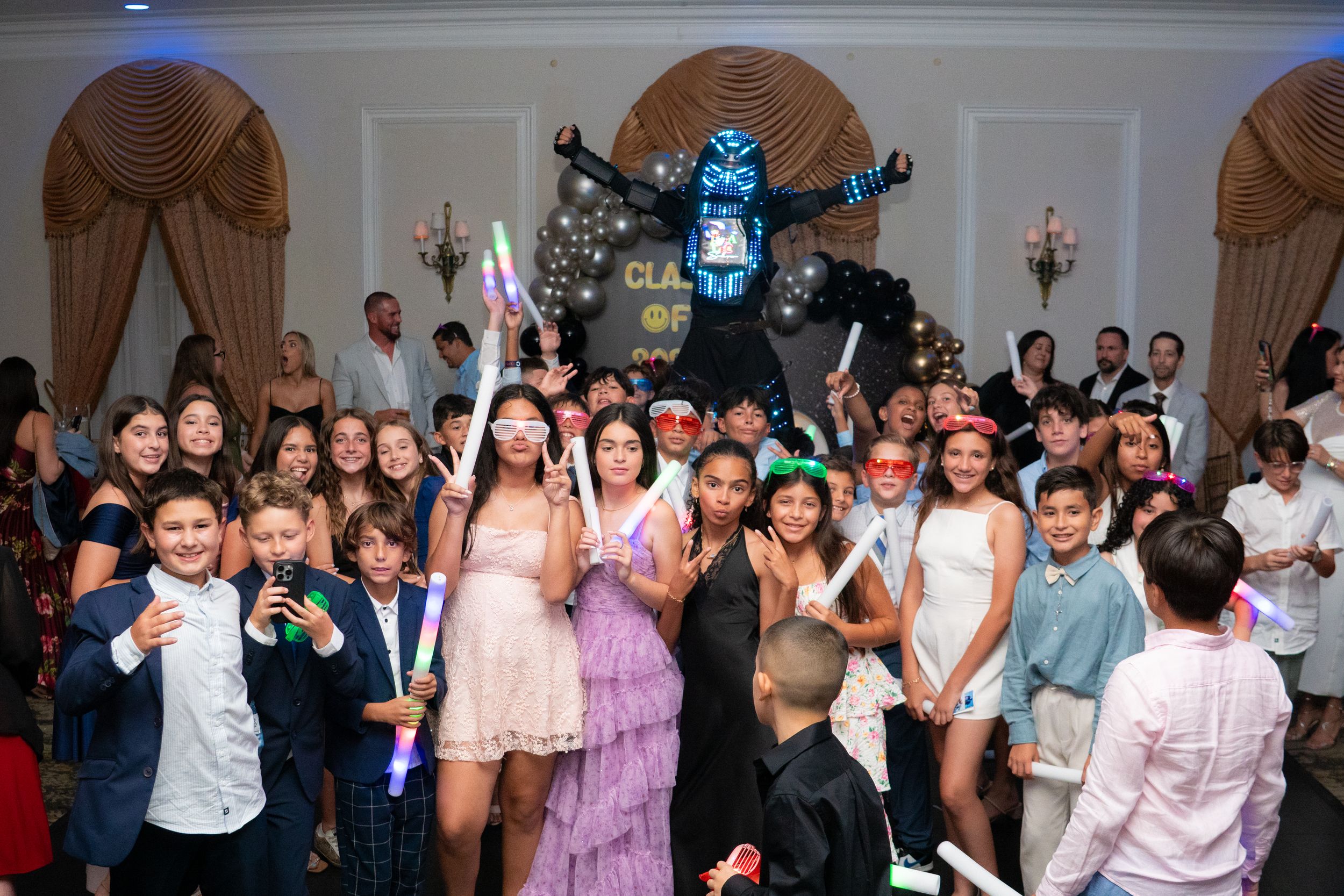 Large group of teens celebrating at a party with glow sticks and fun props in an elegant ballroom.