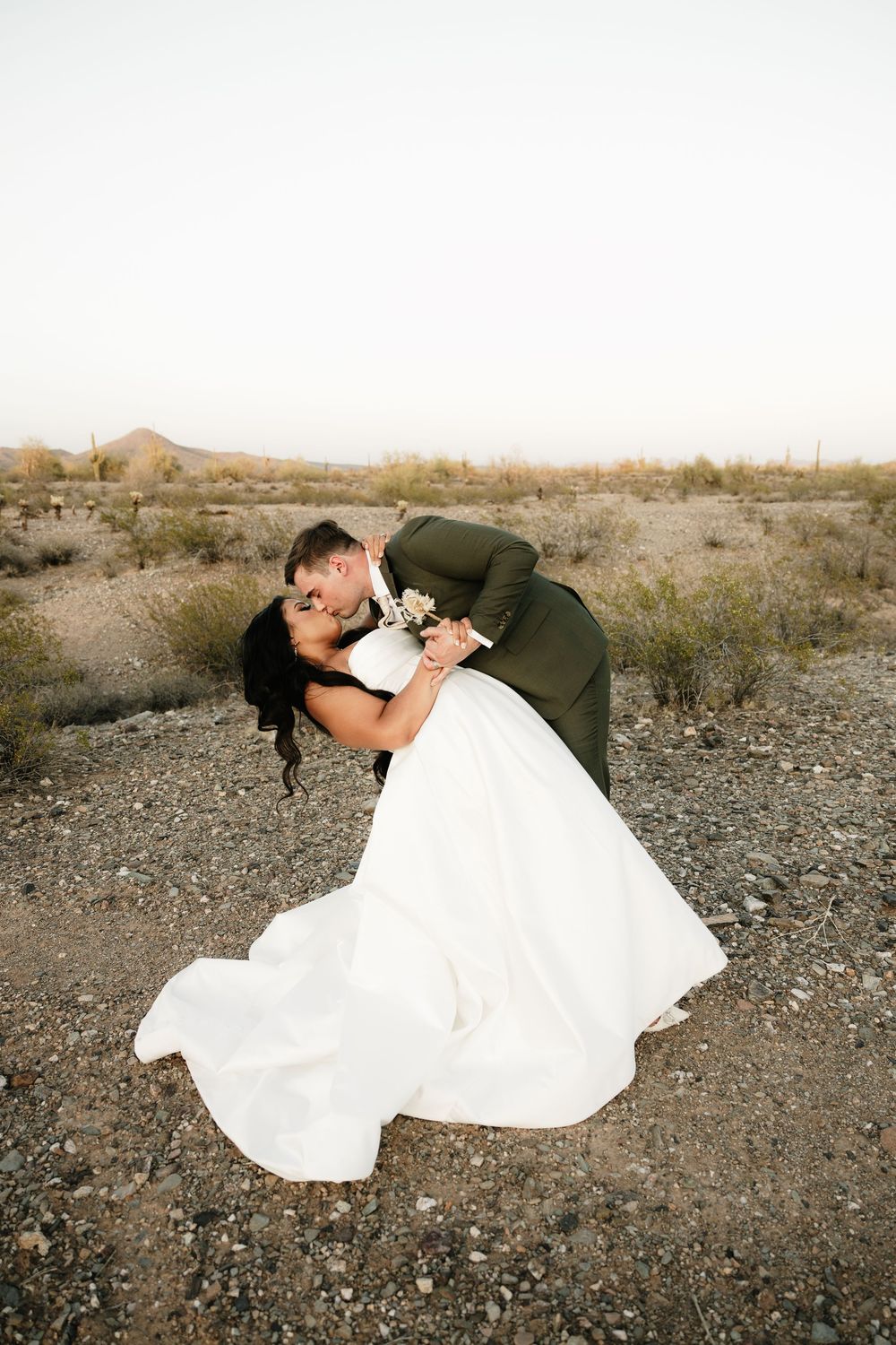 Wedding couple poses dramatically on desert ground with mountains in the distance.