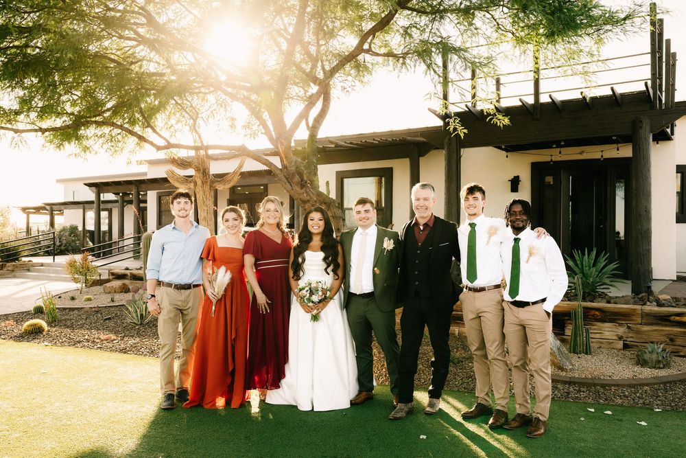 Wedding party group photo on lawn with desert landscape and buildings in background.
