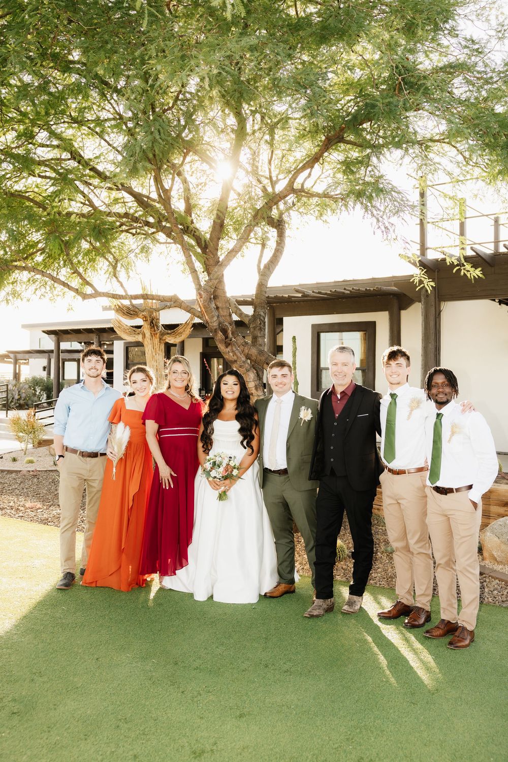 Wedding party in red and khaki attire poses under shady trees on green lawn.