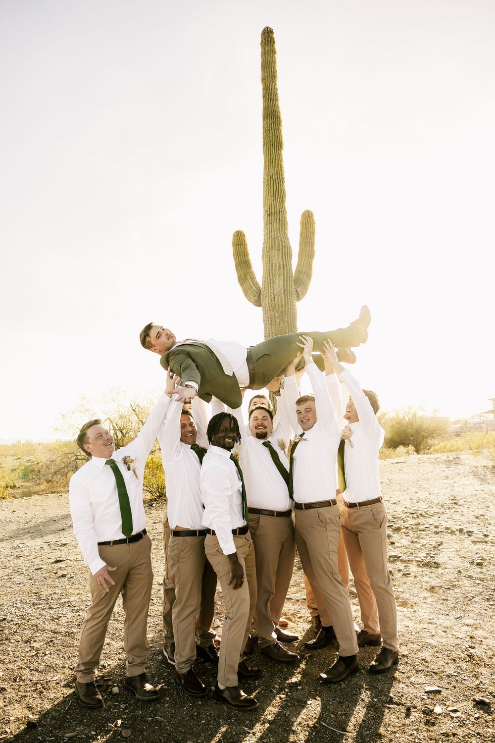 Groomsmen in khaki pants and white shirts pose playfully by desert cactus at sunset.
