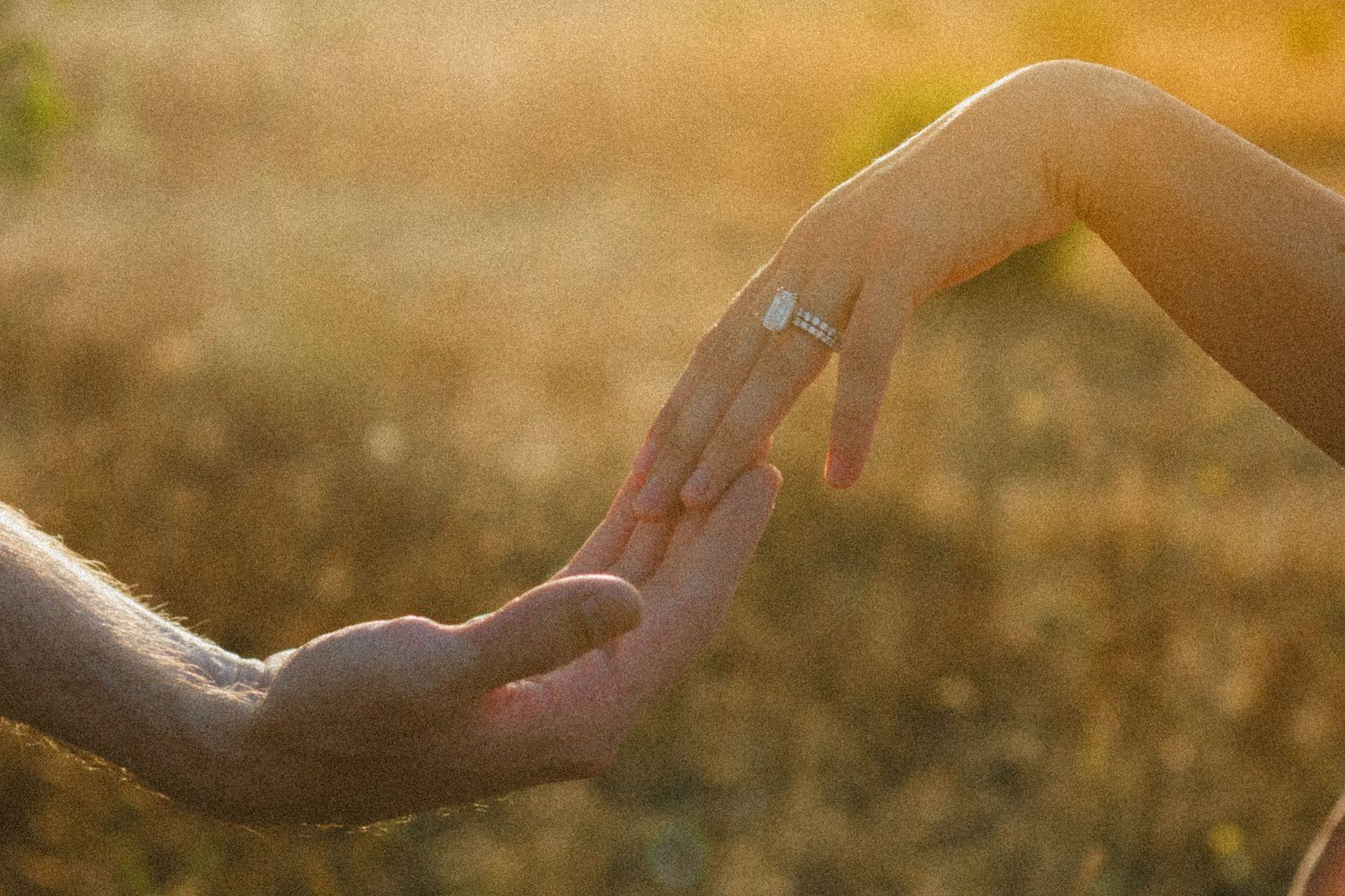 Hands reaching towards each other in warm golden sunlight with an engagement ring glinting.