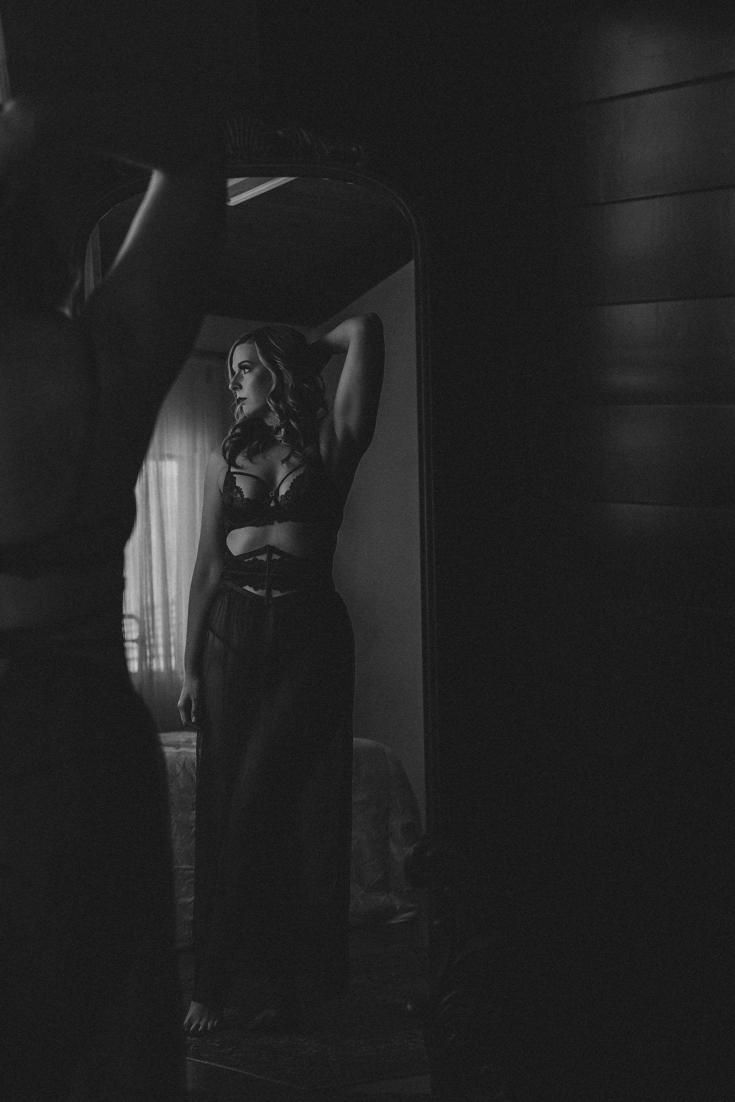 Moody black and white boudoir photo with dramatic window lighting and elegant pose.