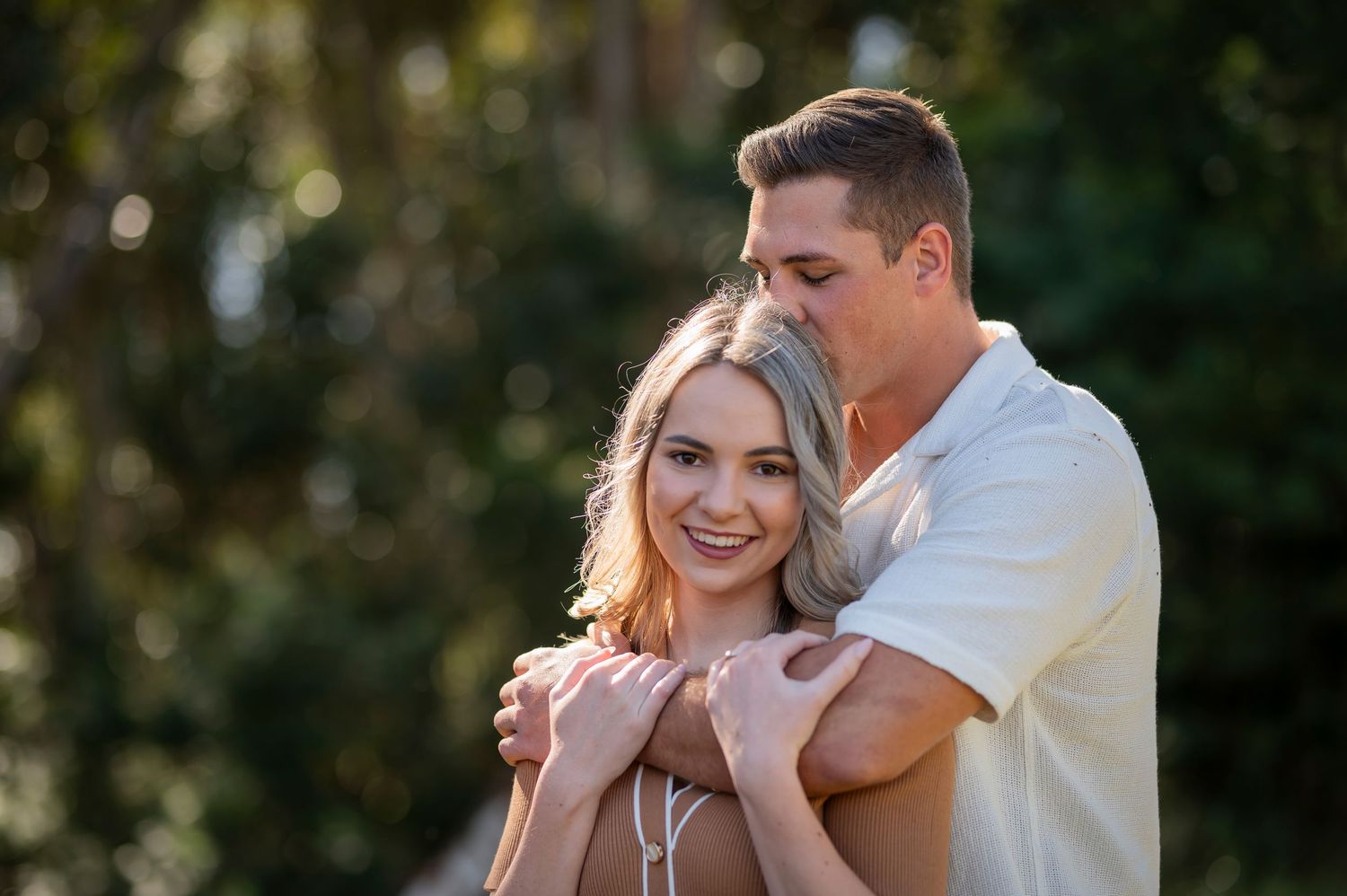 A romantic outdoor portrait shows a couple embracing in warm evening sunlight with a natural bokeh background.