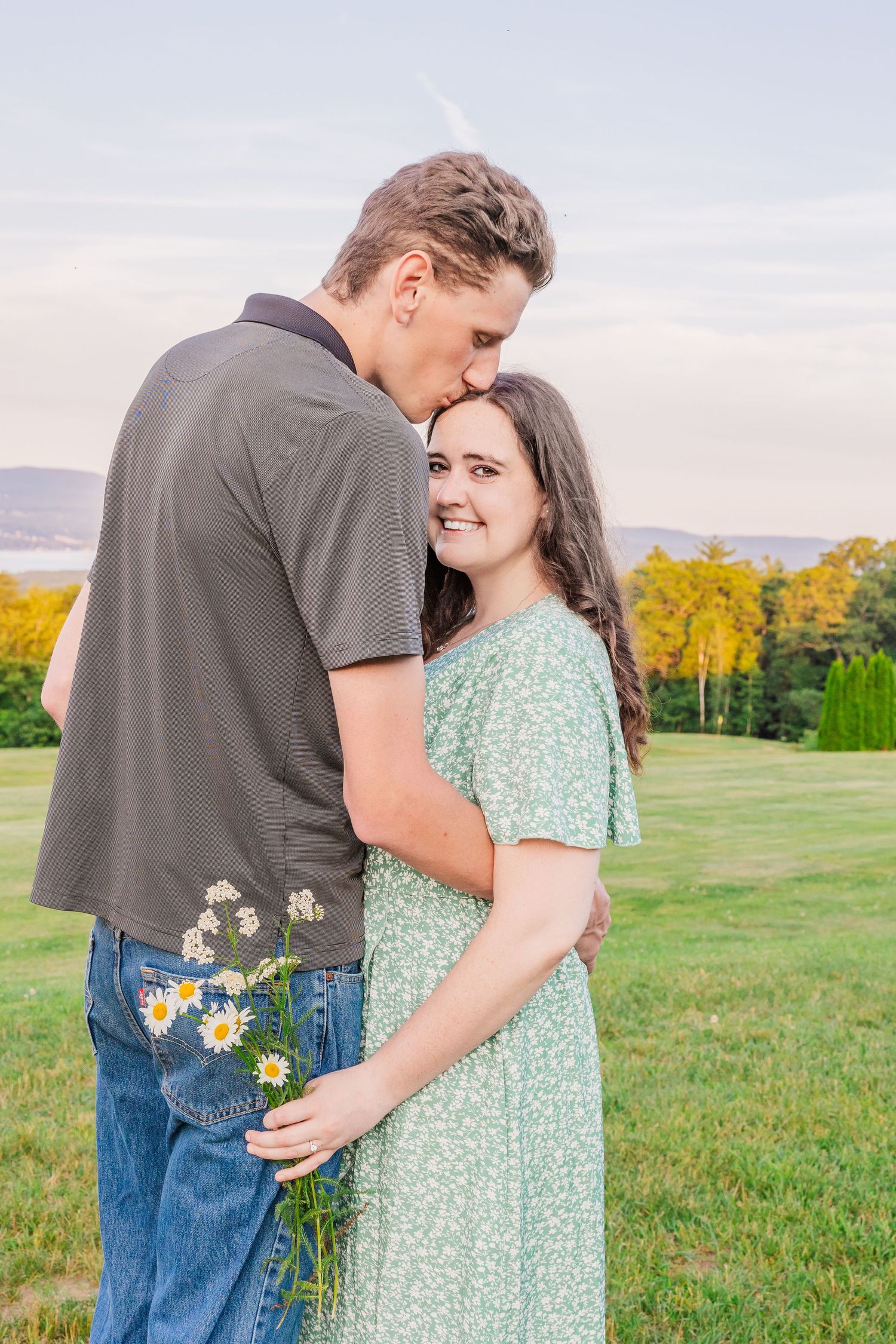 A romantic couple embraces in a field at sunset with mountains and autumn foliage in the background.