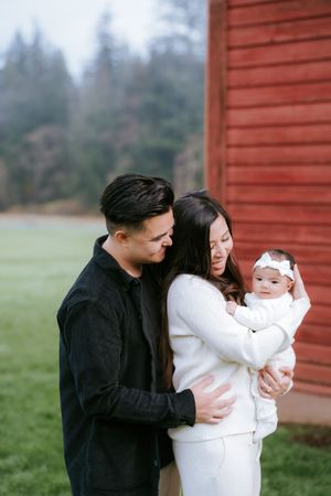 Family shares tender moment together with baby outdoors by red barn wall on misty day.