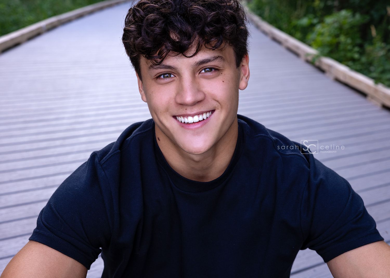 A person wearing a navy blue t-shirt smiles warmly while sitting outdoors on a wooden boardwalk surrounded by greenery.