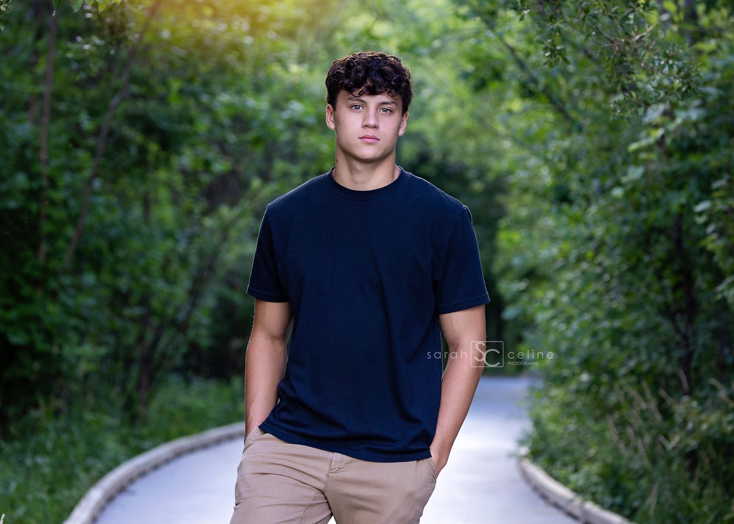 Young person wearing a dark-colored t-shirt and khaki pants standing casually on a curved path surrounded by greenery.