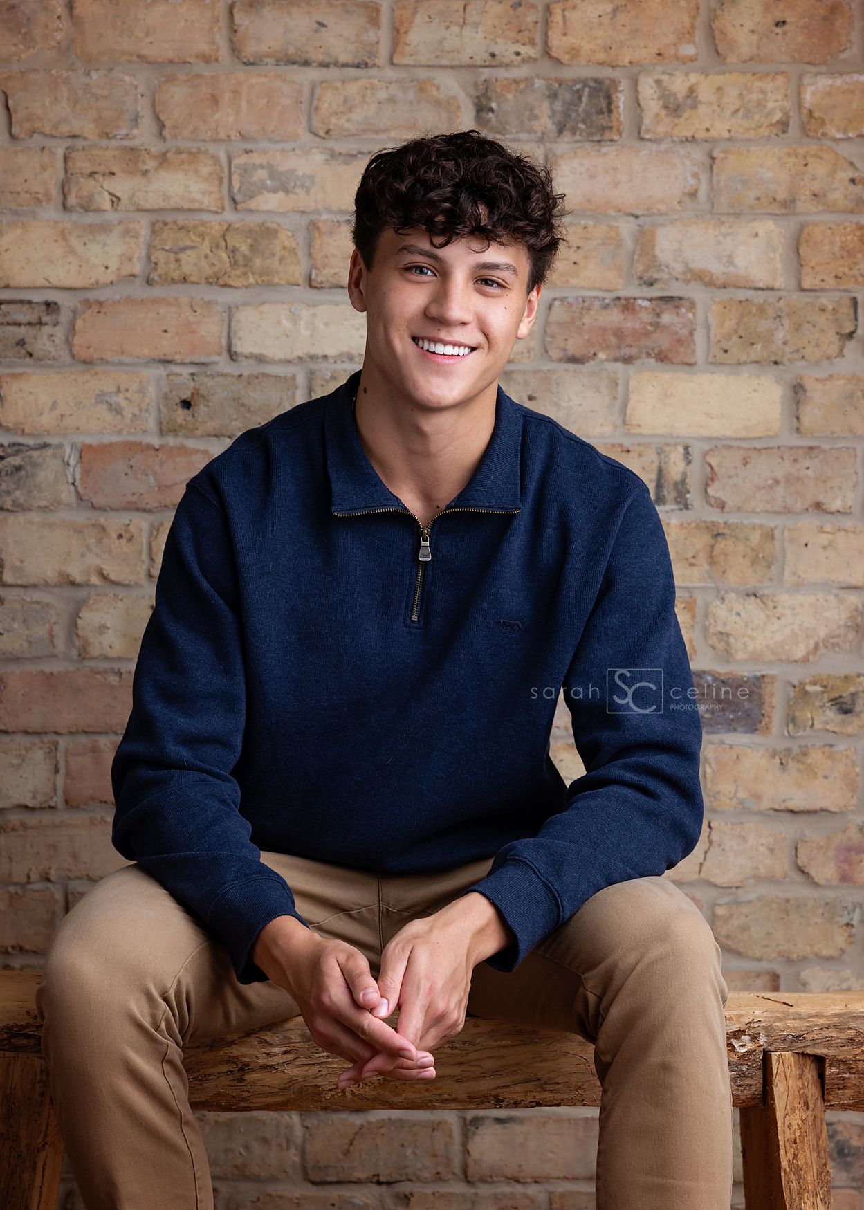 A person wearing a navy blue quarter-zip sweater and khaki pants sits casually on a rustic wooden bench against a brick wall.