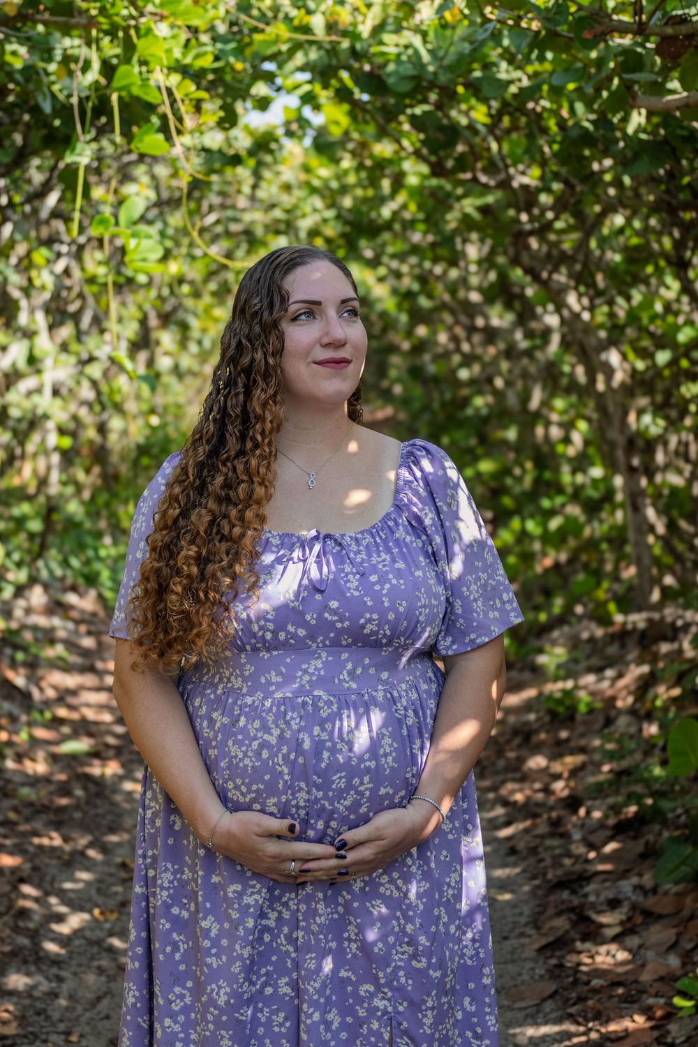 Woman in purple floral dress poses in dappled sunlight among green foliage.