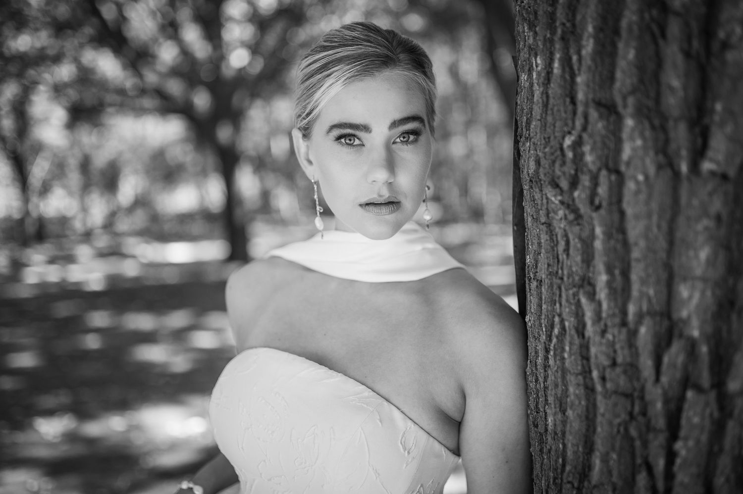 Black and white portrait photo taken outdoors near a tree trunk with dappled sunlight creating a dramatic lighting effect.