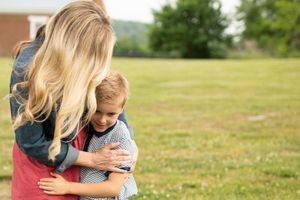 A mother with blonde hair tenderly hugs her young son in a green grassy field outdoors.