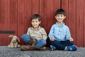 Two young boys wearing cowboy boots standing in front of a red barn with a toy dog outside in Peachtree City