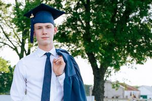 Young male graduate in cap and gown holding diploma outdoors with green trees in background.
