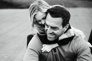 Black and white photo of a happy couple embracing outdoors, woman hugging man from behind, both smiling joyfully.