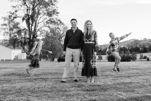 Black and white family photo of couple with two children jumping joyfully in a grassy field with trees in background.