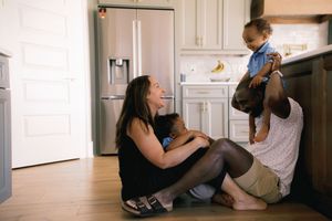 Family sitting on kitchen floor, mother holding baby while father lifts toddler in bright modern home.