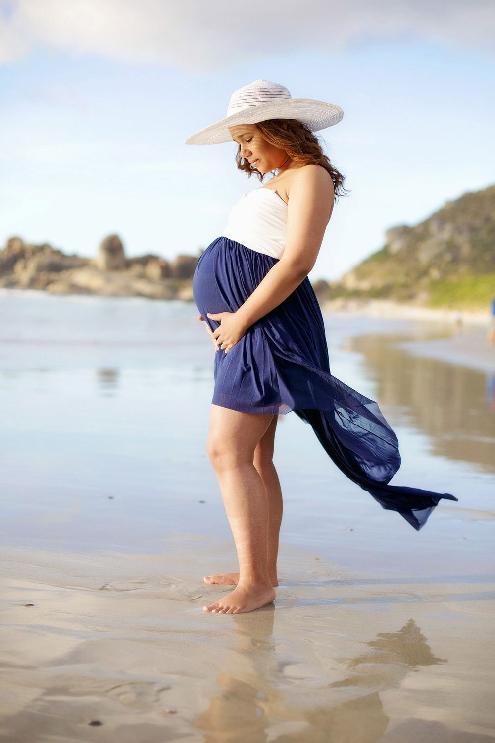 Pregnant woman with flowing dress at beach, admiring belly, Melbourne shoot