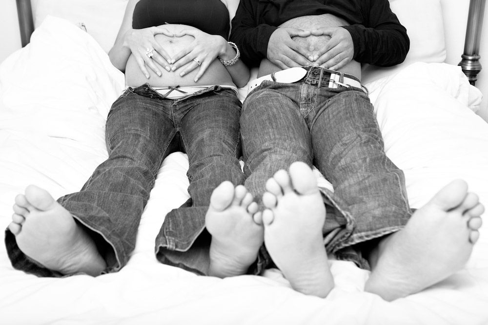 A pregnant woman and her partner lie on a bed, forming a heart shape with their hands over her baby bump creating a tender and loving moment